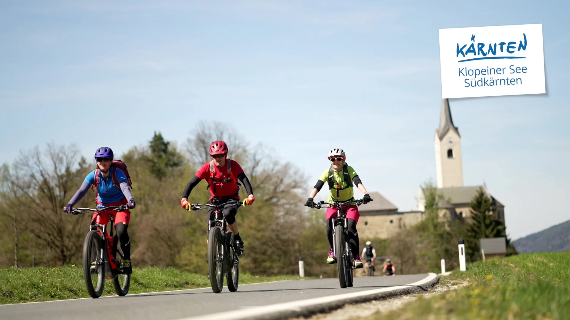 Drei Radfahrerinnen und Radfahrer auf einer Landstraße vor einer Kirche in Südkärnten