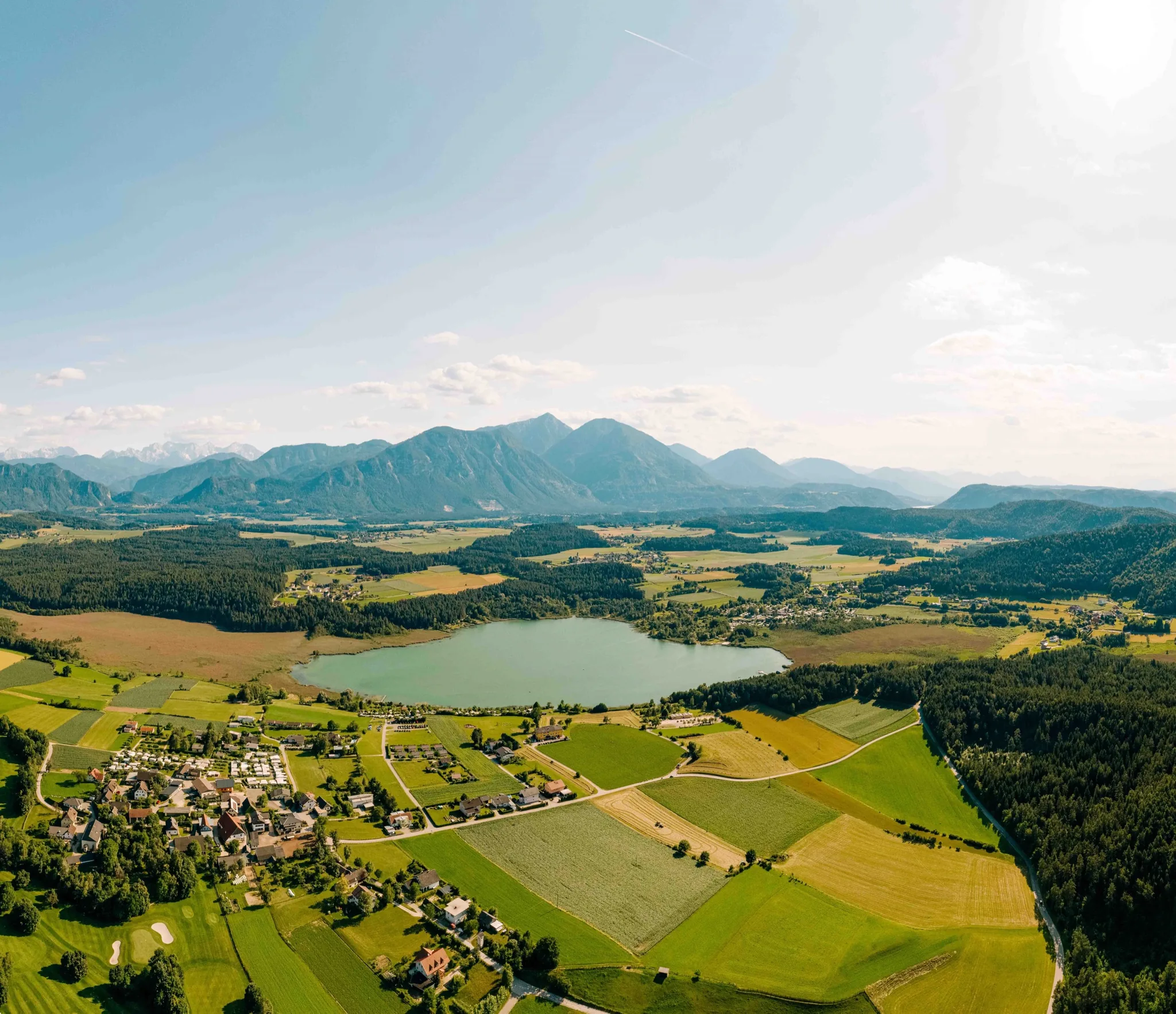Der Turnersee aus der Luft, eingebettet zwischen Feldern und Wald, dahinter die Karawanken