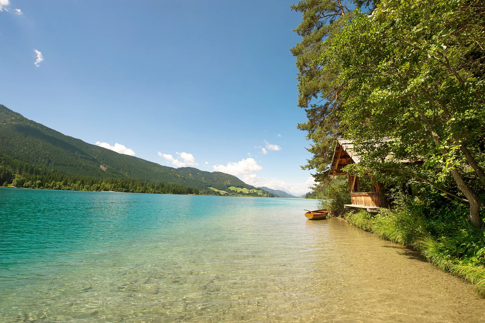 Flaches türkises Wasser am Ufer des Weissensees, daneben ein Bootshaus